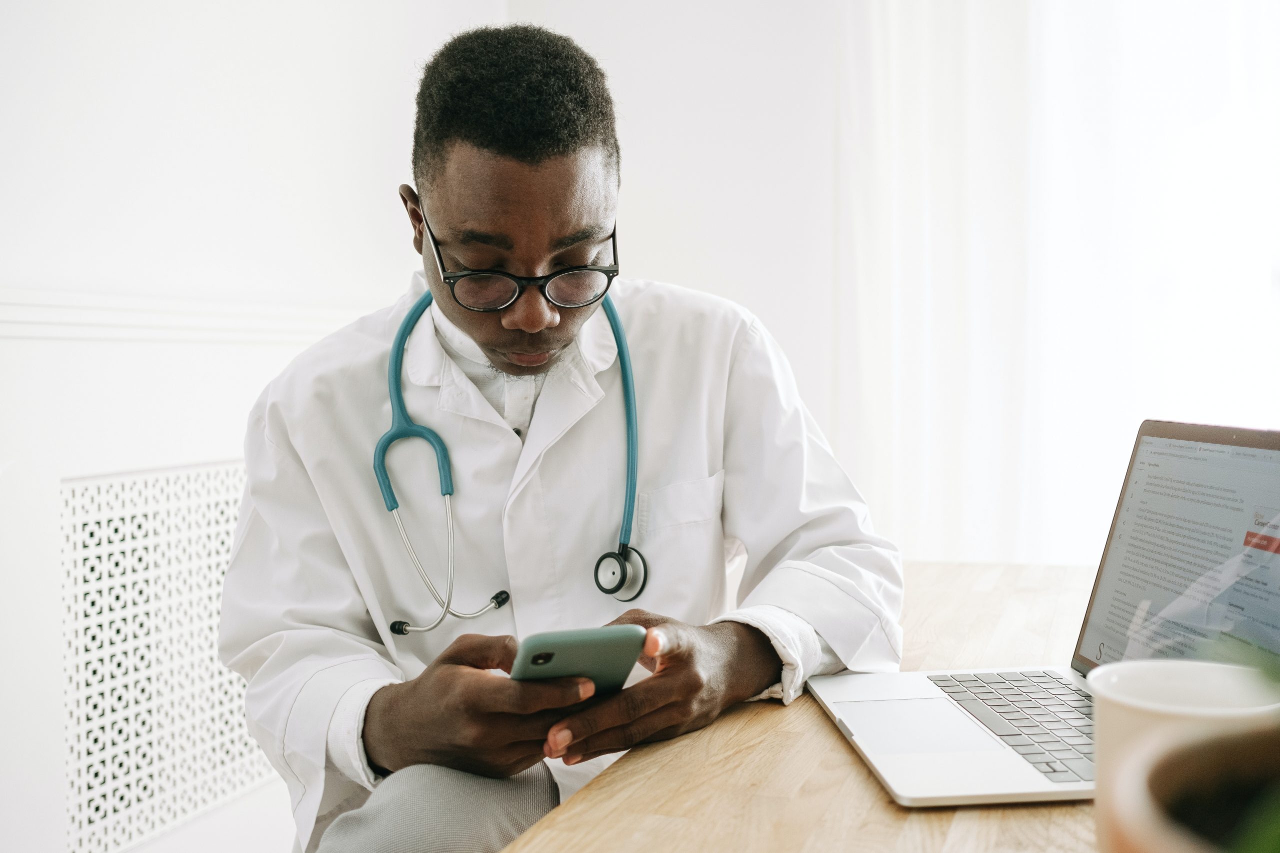 African American male doctor with serious expression on cell phone sitting at desk with computer